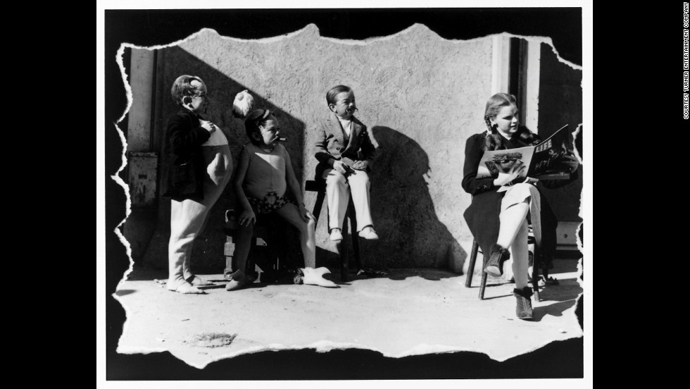 Judy Garland reads a copy of Life magazine on set as three of the actors playing Munchkins stand by.